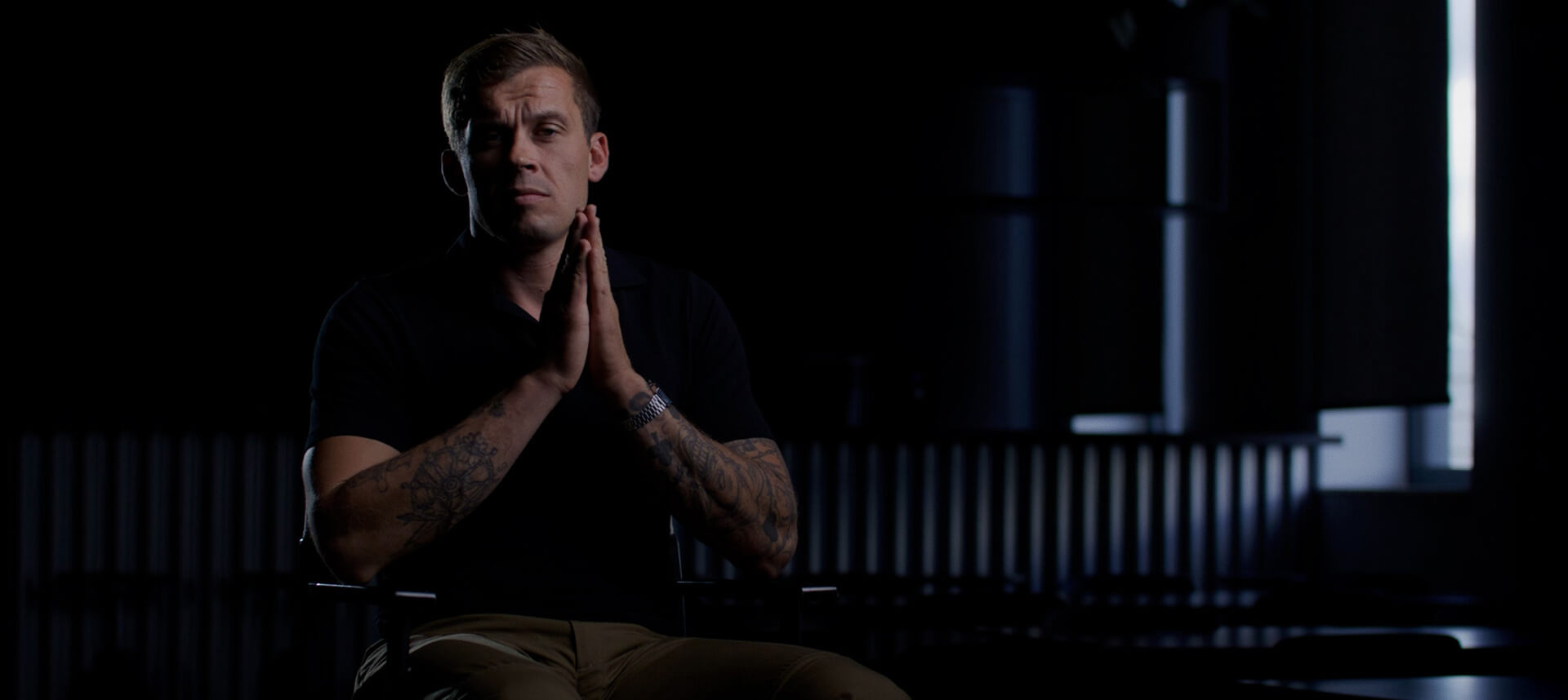 Man seated in a dark room with hands clasped, appearing reflective and focused, symbolizing contemplation of situational awareness.